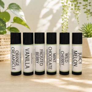 product line of 6 lip balms that consist of Vanilla, Mint Chocolate, Orange Creamsicle, Frosted Berry, Wild Cherry, and Juicy Melon, all in a bright room with leafy plants in the background.